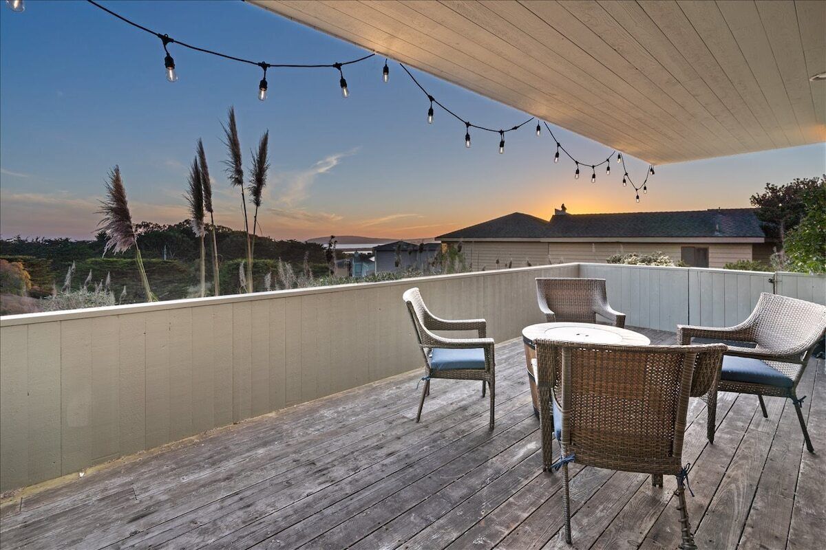 A wooden deck with wicker furniture and string lights, overlooking a landscape with a sunset.