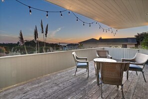 Una terraza de madera con muebles de mimbre y luces colgantes, con vista a un paisaje al atardecer.