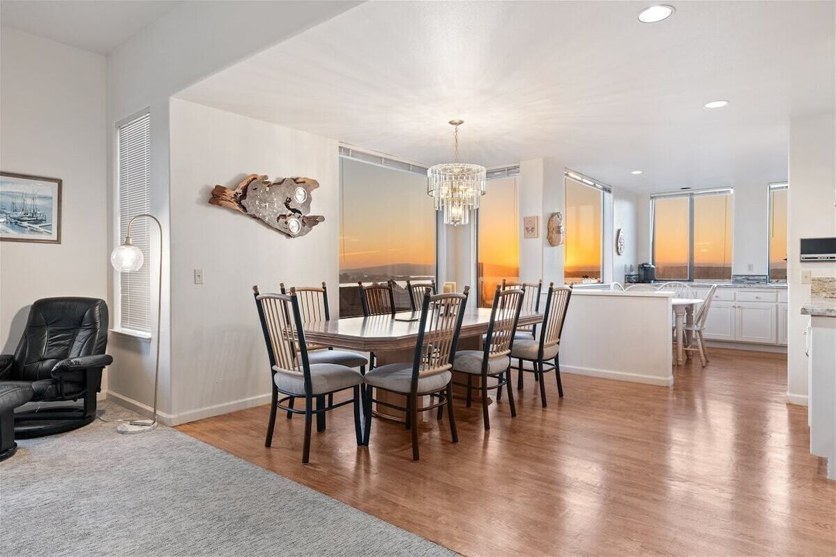 Ein moderner Essbereich mit Holztisch und Stühlen, einer Chandelierleuchtre und Blick auf den Sonnenuntergang durch große Fenster.