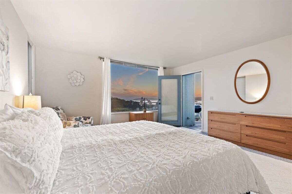 A bedroom with a large bed, a wooden dresser, a round mirror, and a view of the outside through a window.