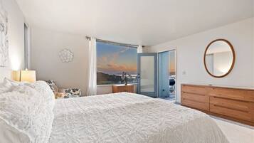 A bedroom with a large bed, a wooden dresser, a round mirror, and a view of the outside through a window.