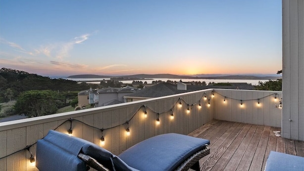 Un balcone con luci a tendina, vista sul tramonto sull'acqua e un paesaggio in lontananza.