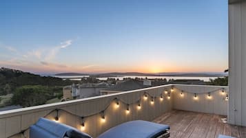 A balcony with string lights, a view of the sunset over water, and a distant landscape.