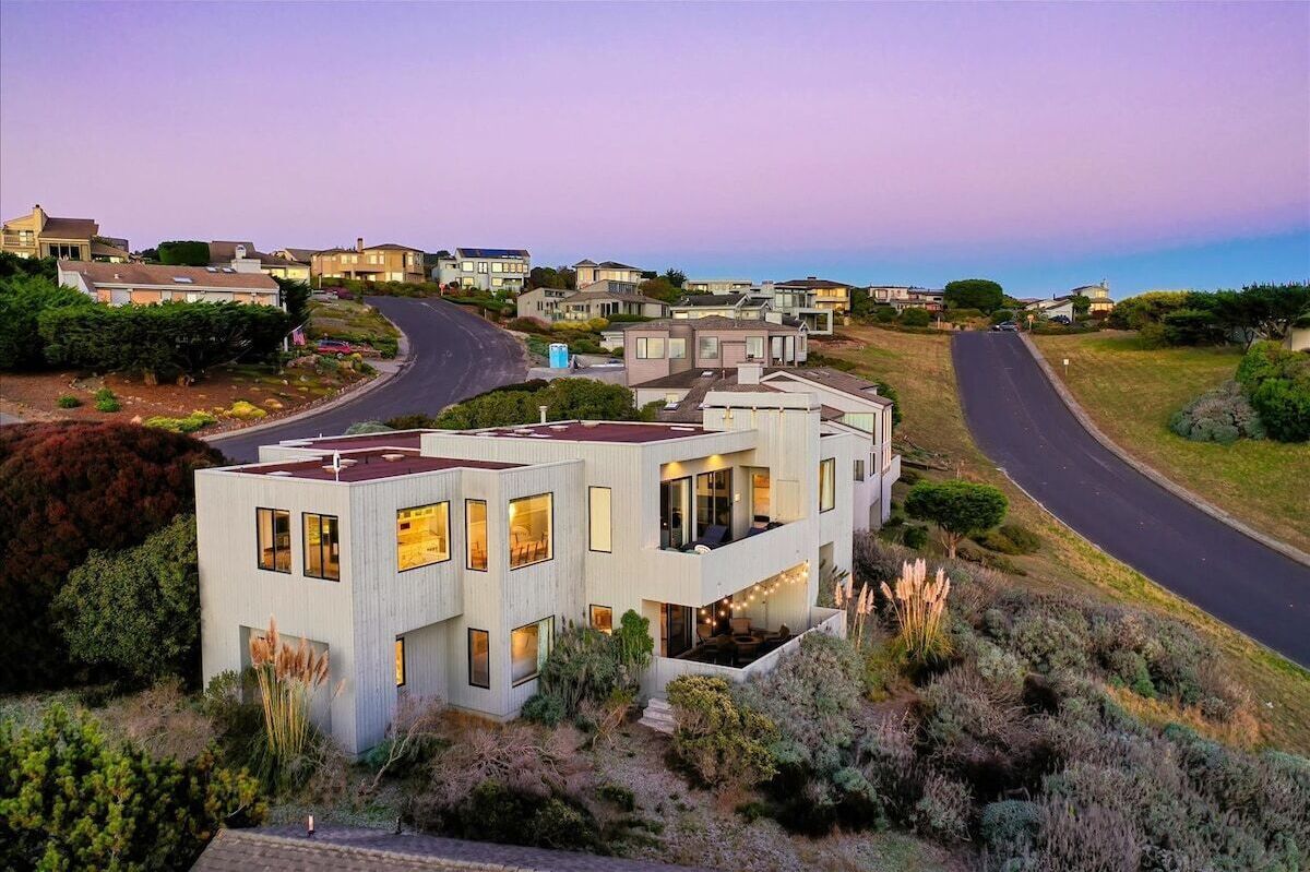 A modern two-story house with a flat roof, large windows, and a visible outdoor seating area.