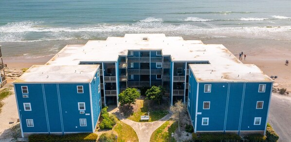 A beachfront hotel with a central building and two wings, featuring balconies and a view of the ocean.