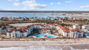 Un resort frente a la playa con varias construcciones, una piscina central y una playa en primer plano.