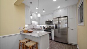 A modern kitchen with white cabinets, a stainless steel refrigerator, a microwave, and a granite countertop. There are two wooden stools at the island. - Shipwatch 1411 - Point Unit (North Topsail Beach)