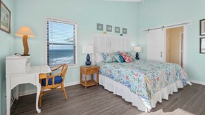 A bedroom with a bed, a desk, a chair, a window with blinds, and a door. - Shipwatch 1411 - Point Unit (North Topsail Beach)