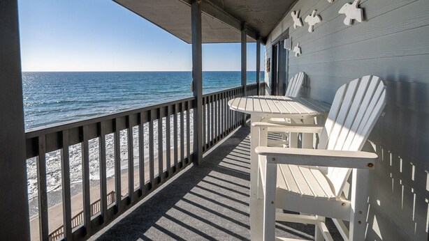 A covered deck with two Adirondack chairs and a table, overlooking a beach with the ocean. - Shipwatch 1411 - Point Unit (North Topsail Beach)