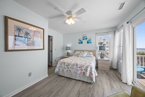 A bedroom with a bed, a ceiling fan, a nightstand with a lamp, a framed picture, and a view of the outdoors. - New River Inlet Rd. 1338 (North Topsail Beach)