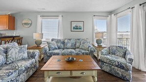 A living room with a wooden coffee table, floral-patterned sofas, a dining area with a wooden table and chairs, and large windows with white curtains. - New River Inlet Rd. 1338 (North Topsail Beach)
