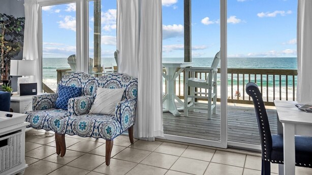 A room with a large sliding glass door leading to a balcony with a view of the ocean. There is a patterned sofa and a small table with chairs. - Anchor of Santorini - 5 Bedroom Oceanfront Townhouse Sleeps 15 (Surf City)