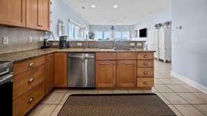 A modern kitchen with wooden cabinets, a stainless steel dishwasher, and a granite countertop. - Anchor of Santorini - 5 Bedroom Oceanfront Townhouse Sleeps 15 (Surf City)