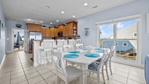 A modern kitchen with wooden cabinets, a stainless steel refrigerator, and a dining area with white chairs and blue placemats. - Anchor of Santorini - 5 Bedroom Oceanfront Townhouse Sleeps 15 (Surf City)