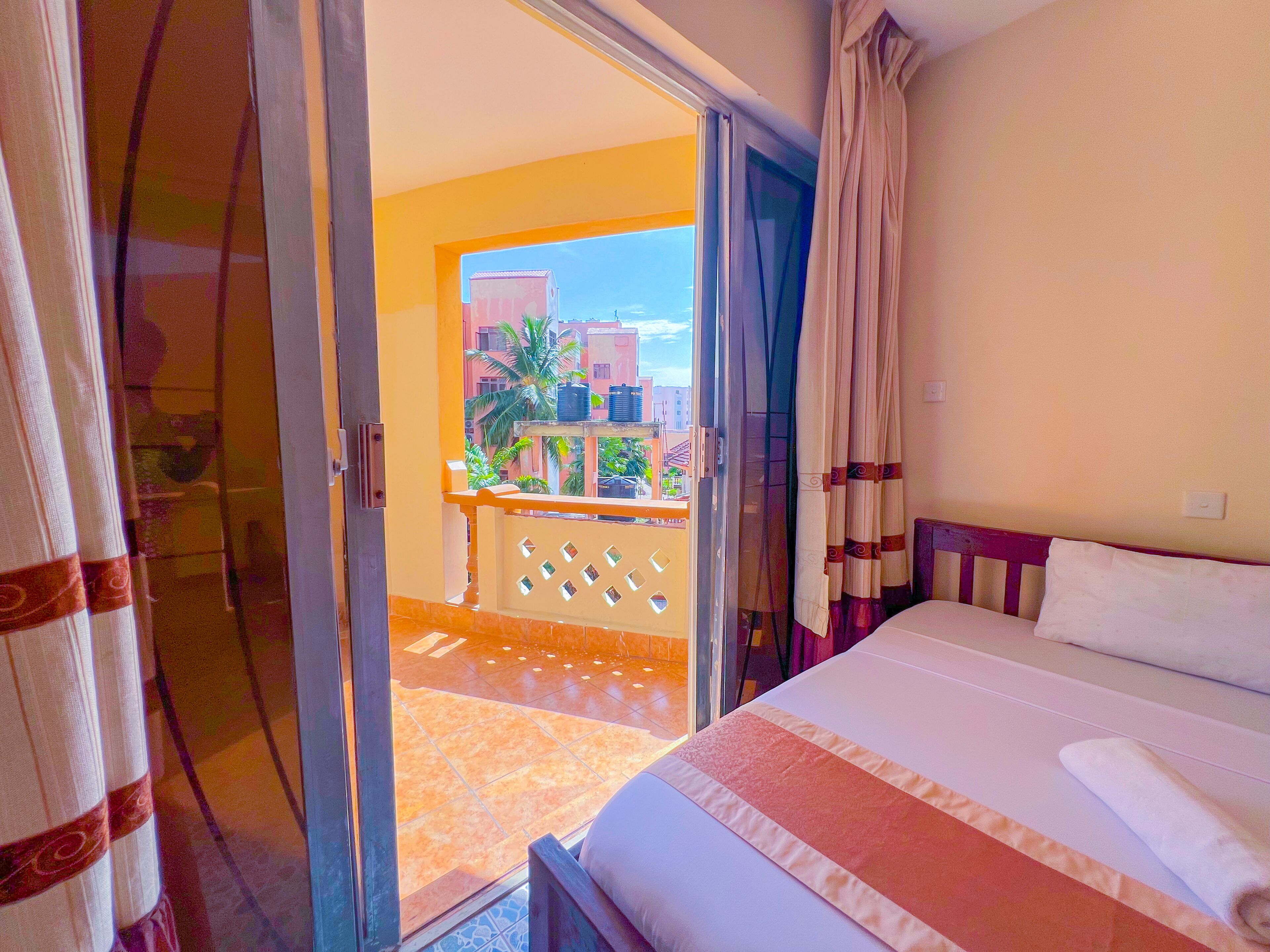 Quarto de hotel com cama, varanda e vista para edifícios e palmeiras.