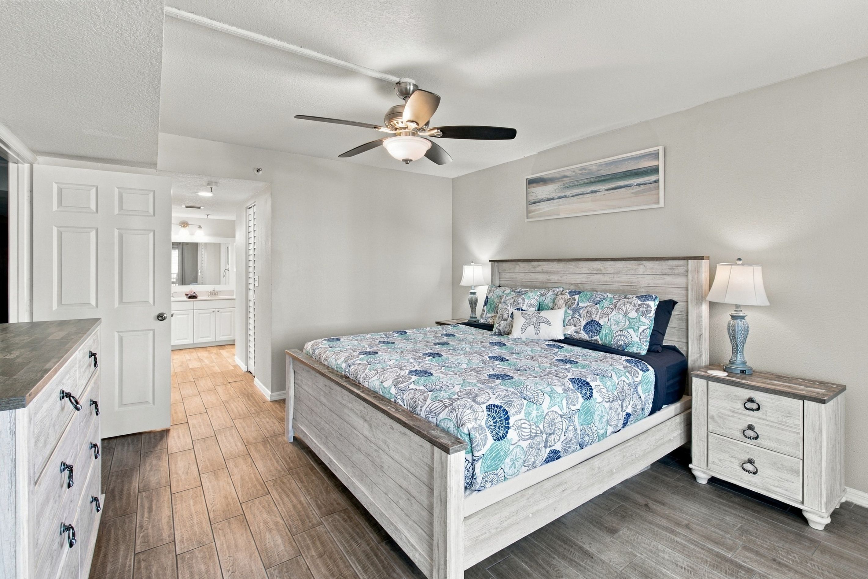 Une chambre à coucher avec un lit, des tables de chevet, un ventilateur de plafond, un tableau et une salle de bain visible à travers une porte ouverte.