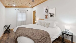 Quarto com teto de madeira, cama com roupa de cama branca, manta marrom, ventilador de teto e duas luminárias fixadas na parede.