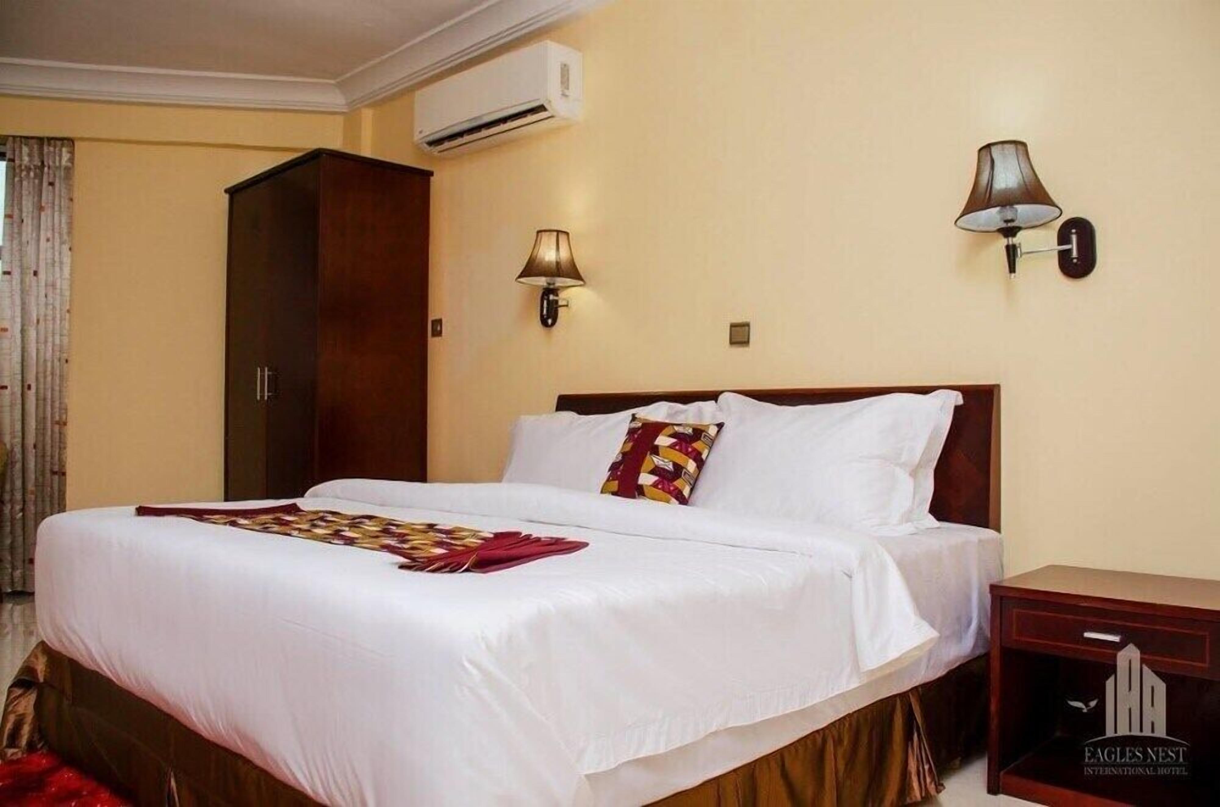 Habitación de hotel con una cama grande, dos lámparas fijadas a la pared, un equipo de aire acondicionado y una mesita de noche de madera con un logotipo.
