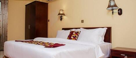 Habitación de hotel con una cama grande, dos lámparas fijadas a la pared, un equipo de aire acondicionado y una mesita de noche de madera con un logotipo.