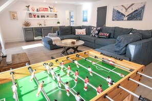 A living room with a foosball table, a grey sofa, and a kitchen area in the background. - Luxury Waterfront Home w/ Heated Pool and Hot Tub (Yarmouth)