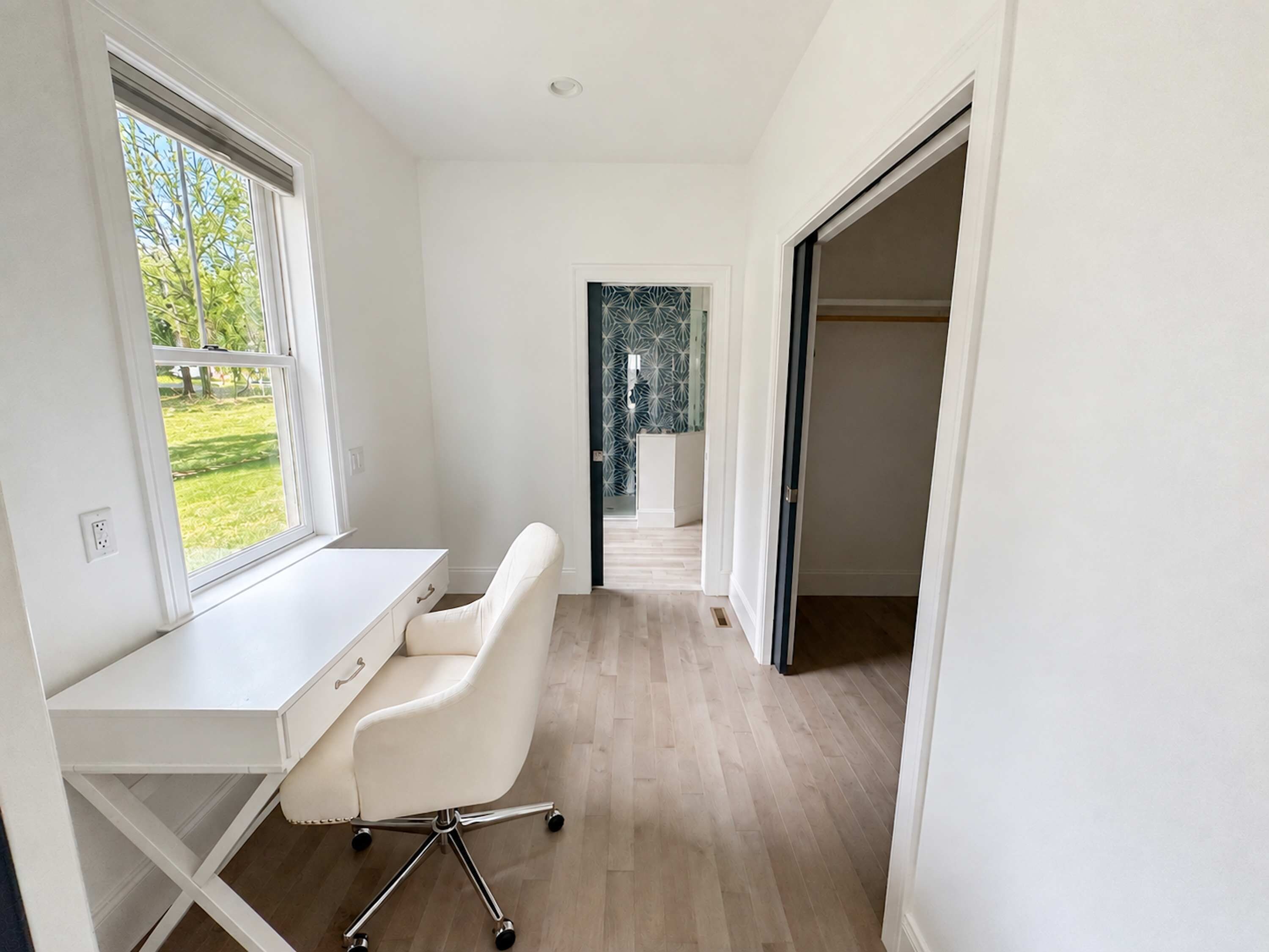 A modern, minimalistic room with a white desk, a leather chair, and a view of a garden through a window.