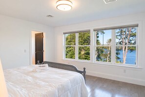 A hotel room with a bed, a window with a view of trees and water, and a door leading to another room.