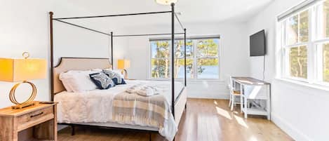A four-poster bed with a canopy, bedside lamps, a wooden nightstand, a desk with a chair, and a television on the wall.