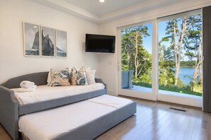 Ein modernes Schlafzimmer mit grauem Schlafsofa, einem Flachbildfernseher und einem großen Fenster mit Blick auf Bäume und Wasser.