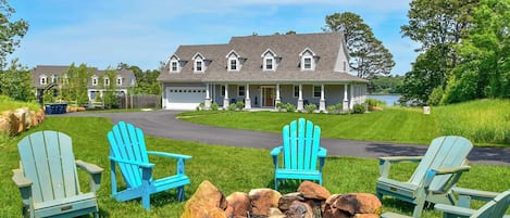 Une grande maison dotée d’un porche, d’un foyer extérieur et de trois fauteuils Adirondack.