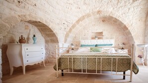 Een slaapkamer met een wit metalen bed, een beddengoed met groene patronen, een witte dressoir met een blauwe vaas, en een witte nachtkastje.