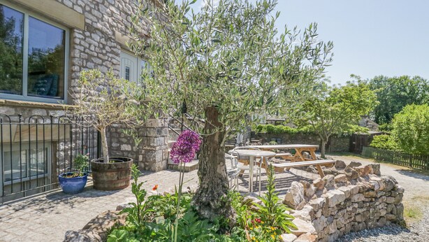 A stone building with a garden featuring a tree, potted plants, and a wooden table and chairs. - Old Watermill (Grange-over-Sands)