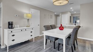 A dining area with a white table, striped chairs, a red vase, a coffee maker, and a lamp on a white dresser.