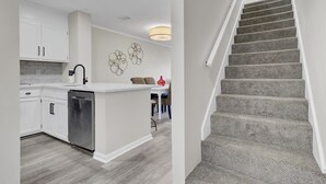 A modern kitchen with white cabinetry, a stainless steel dishwasher, and a dining area with a red vase and two chairs.