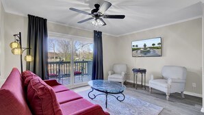 A living room with a red sofa, two armchairs, a glass coffee table, a ceiling fan, and a large window with curtains.