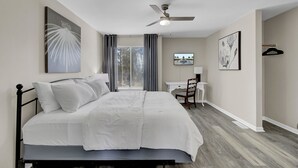 A bedroom with a bed, a desk, a chair, a ceiling fan, and artwork on the walls.