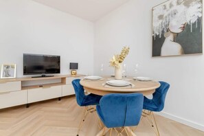 Un espace repas moderne avec une table ronde pour quatre personnes, des chaises en velours bleu, un téléviseur à écran plat posé sur un meuble blanc et une œuvre d’art encadrée accrochée au mur.