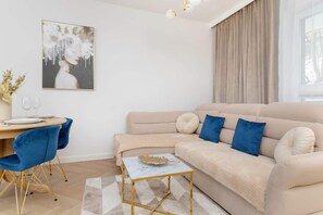 Un salon moderne avec un canapé beige, des fauteuils bleus, une petite table basse et une table à manger avec des chaises. Un tableau orne le mur et une fenêtre est agrémentée de rideaux.