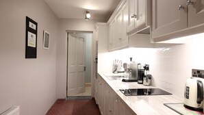 A narrow kitchen with white cabinets, a stainless steel sink, and a coffee machine. - Ryans Roundstone Retreats Quayside Luxury Apt (Roundstone)