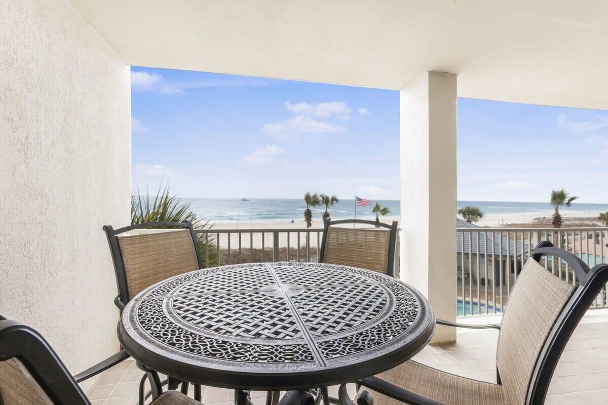 A round table with a lattice design, surrounded by chairs with woven backs, on a balcony with a view of the beach and ocean.