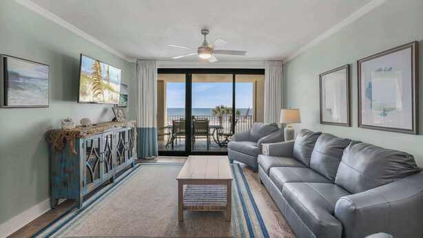 A living room with a grey sofa, a wooden coffee table, and a blue cabinet. There are framed pictures on the walls and a ceiling fan. - Windward Pointe 304- Pristine, Beachfront, Amenities Galore (Orange Beach)