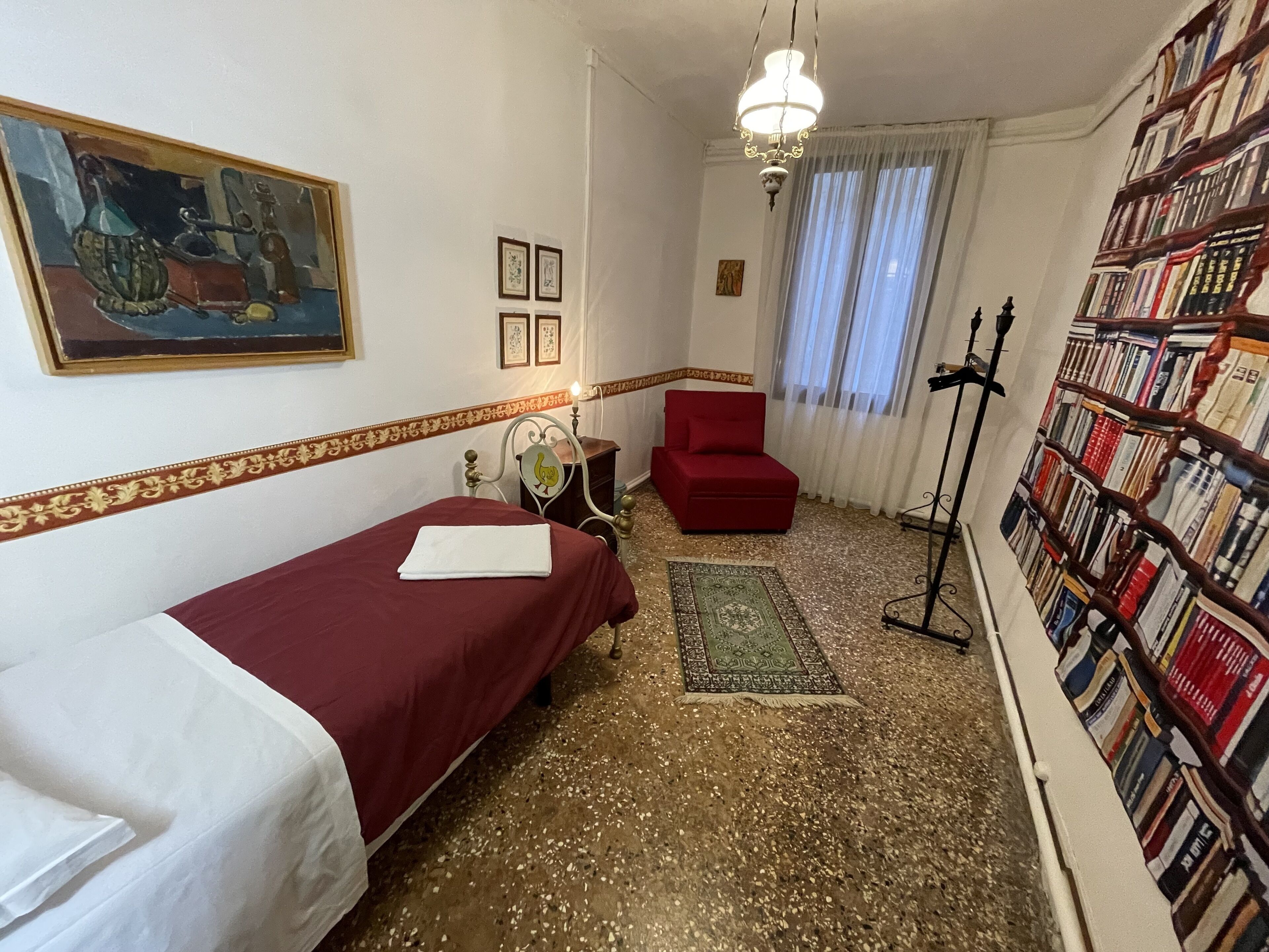 A small, cozy room with a single bed, a red chair, a bookshelf, and a painting on the wall.
