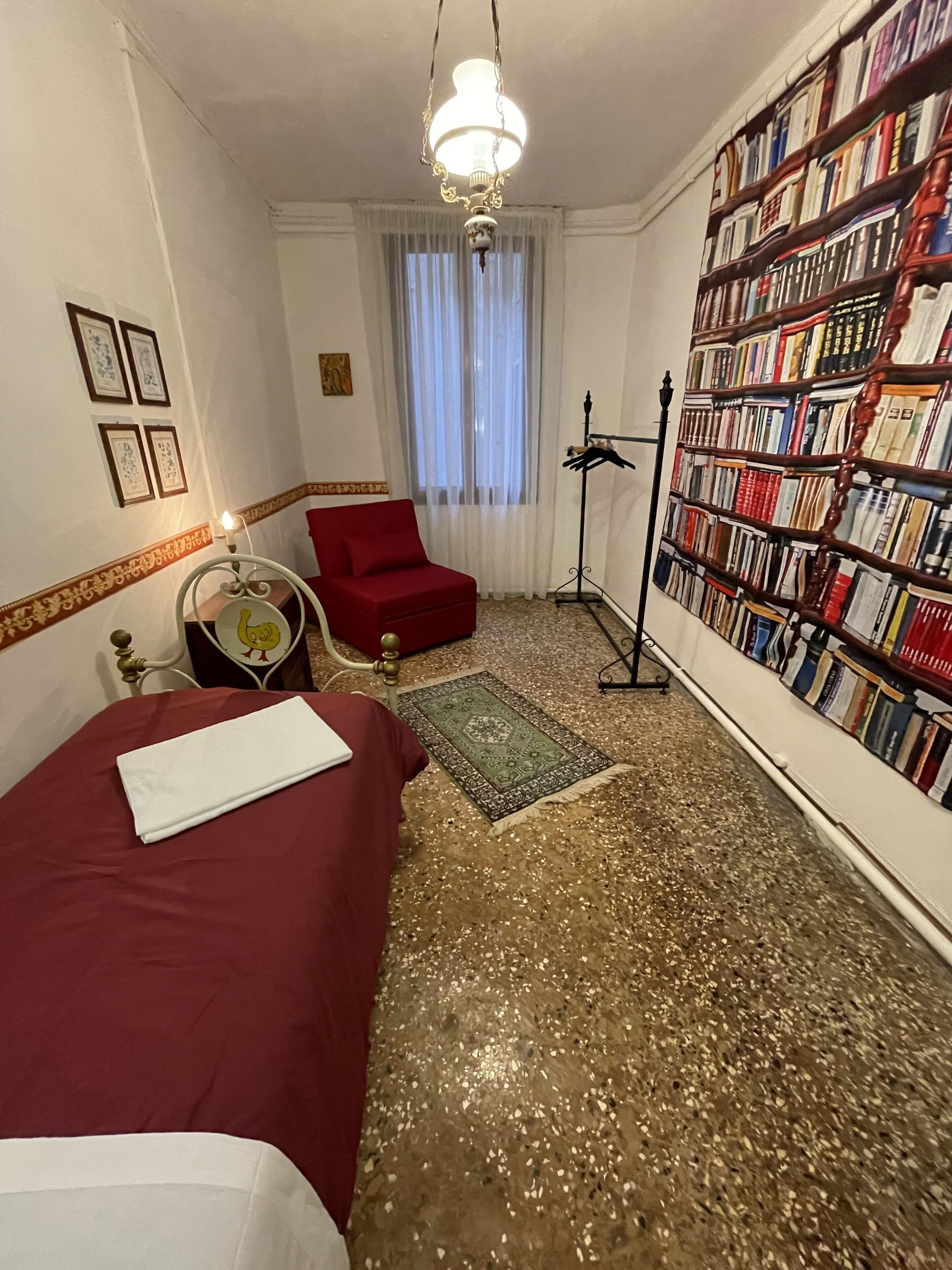 A hotel room with a bed, a red armchair, a bookshelf, and a chandelier.