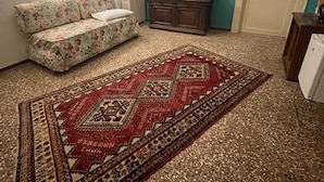 A living room with a floral sofa, a patterned rug, a wooden cabinet, and a radiator. - LIONS VENICE Guest House (Venice)