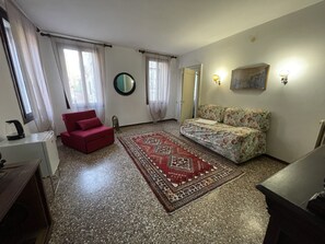 A room with a red chair, a sofa with floral upholstery, a patterned rug, a round mirror, and a painting on the wall. - LIONS VENICE Guest House (Venice)