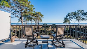 A balcony with two chairs and a small table, overlooking a beach with trees and a clear sky. - 50 Ocean Club-"Ocean Watch" Prime Beachfront Condo (Hilton Head Island)