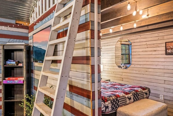 A cozy cabin-style room with a ladder leading to a loft area, a window with a view, and a quilted bed.