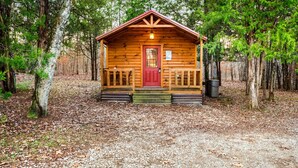 Une petite cabane en bois avec une porte rouge, entourée d’arbres et bordée d’un sol de gravier.
