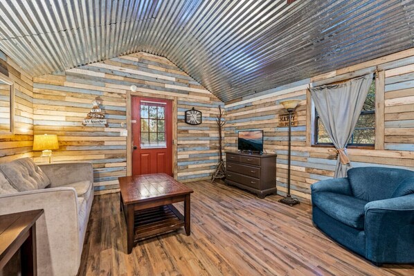 Un interior de cabaña acogedor con piso de madera, una puerta roja, un televisor sobre un aparador, un sofá y una lámpara.