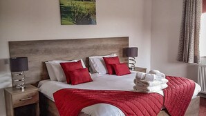 Une chambre à coucher bien rangée, avec une tête de lit en bois, un grand lit recouvert d’une literie rouge et blanche, deux tables de chevet avec des lampes et un tableau accroché au mur.