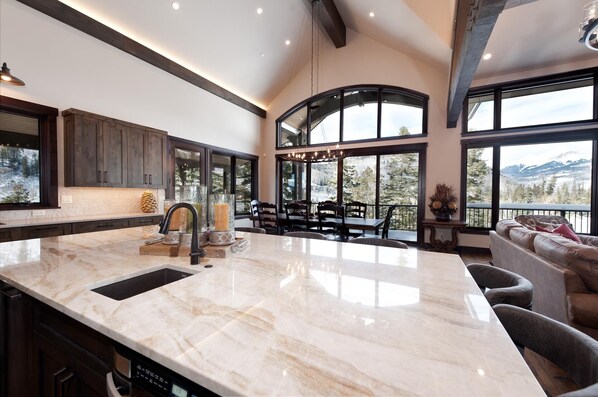 A spacious kitchen with a large marble island, dark wooden cabinets, and a view of a mountainous landscape through large windows.